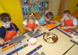 Czworo dzieci siedzi przy stole nakrytym podkładkami z rozłożonymi deskami, w ręku trzymają noże, którymi kroją owoce.